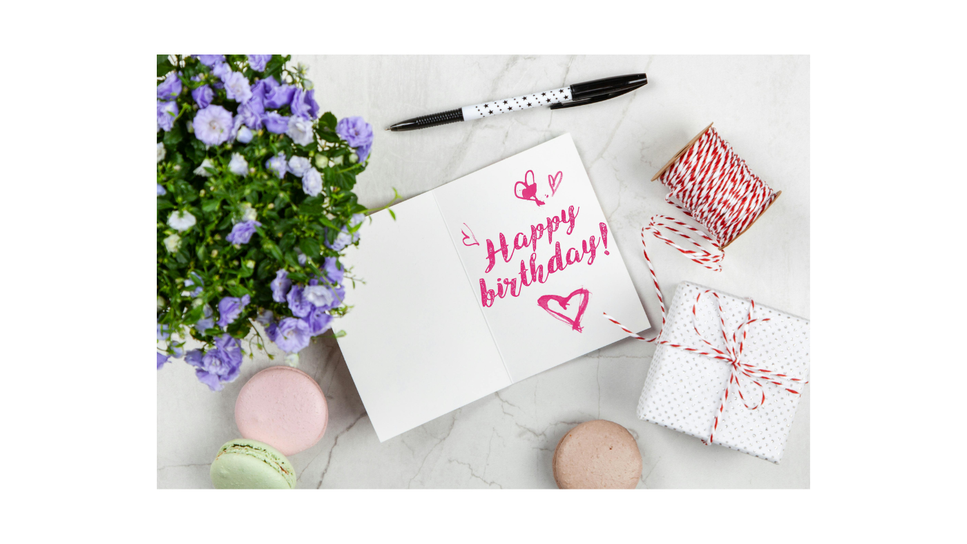 Happy birthday greeting card on a marble table with pastel macarons, purple flowers, a wrapped gift box, and festive twine — perfect birthday celebration scene for unique chocolate gift ideas