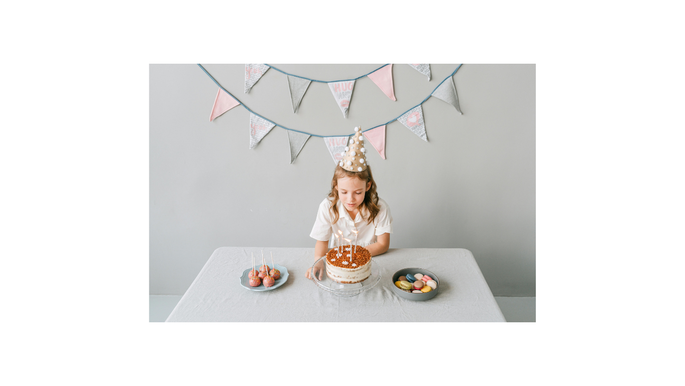 Young girl wearing a birthday hat about to blow out candles on a custom birthday cake, with personalized chocolate treats and macarons on the table, highlighting creative and personalized chocolate gift ideas for birthdays.