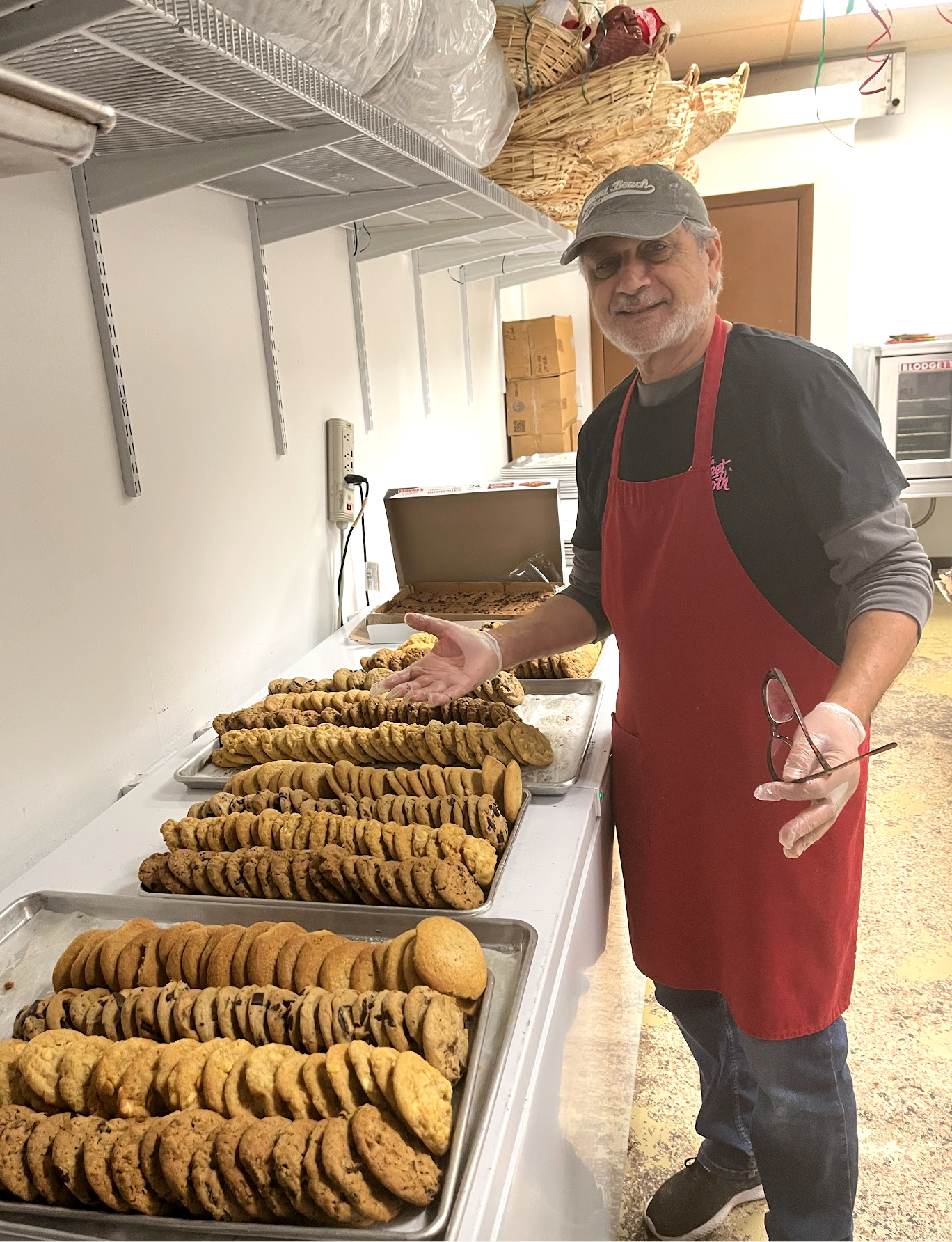 Zalman, expert cookie connoisseur in a red apron and a "key west" t-shirt and cap, standing in the kitchen at The Sweet Tooth next to cooling racks filled with dozens of freshly baked cookies.
