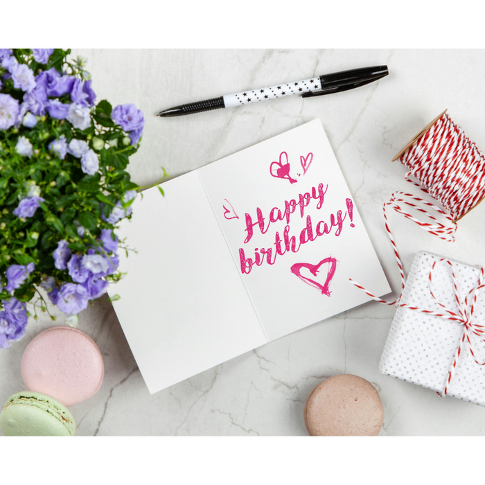 Happy birthday greeting card on a marble table with pastel macarons, purple flowers, a wrapped gift box, and festive twine — perfect birthday celebration scene for unique chocolate gift ideas