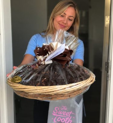 Woman at her front door holding a cellophane-wrapped chocolate basket with a Sweet Tooth gift bag