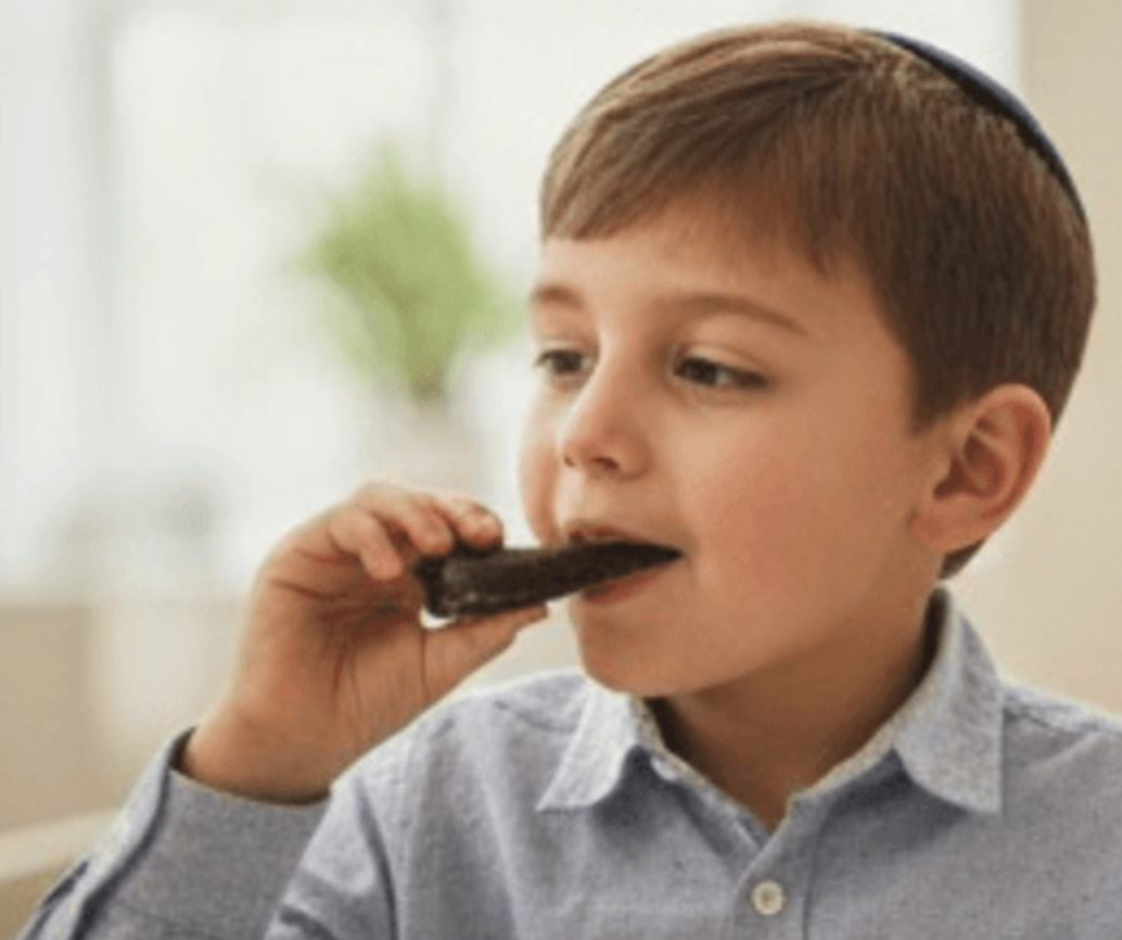 A young boy wearing a blue kippah and a light blue button-down shirt is shown in a close-up profile, eating a dark chocolate shofar from The Sweet Tooth