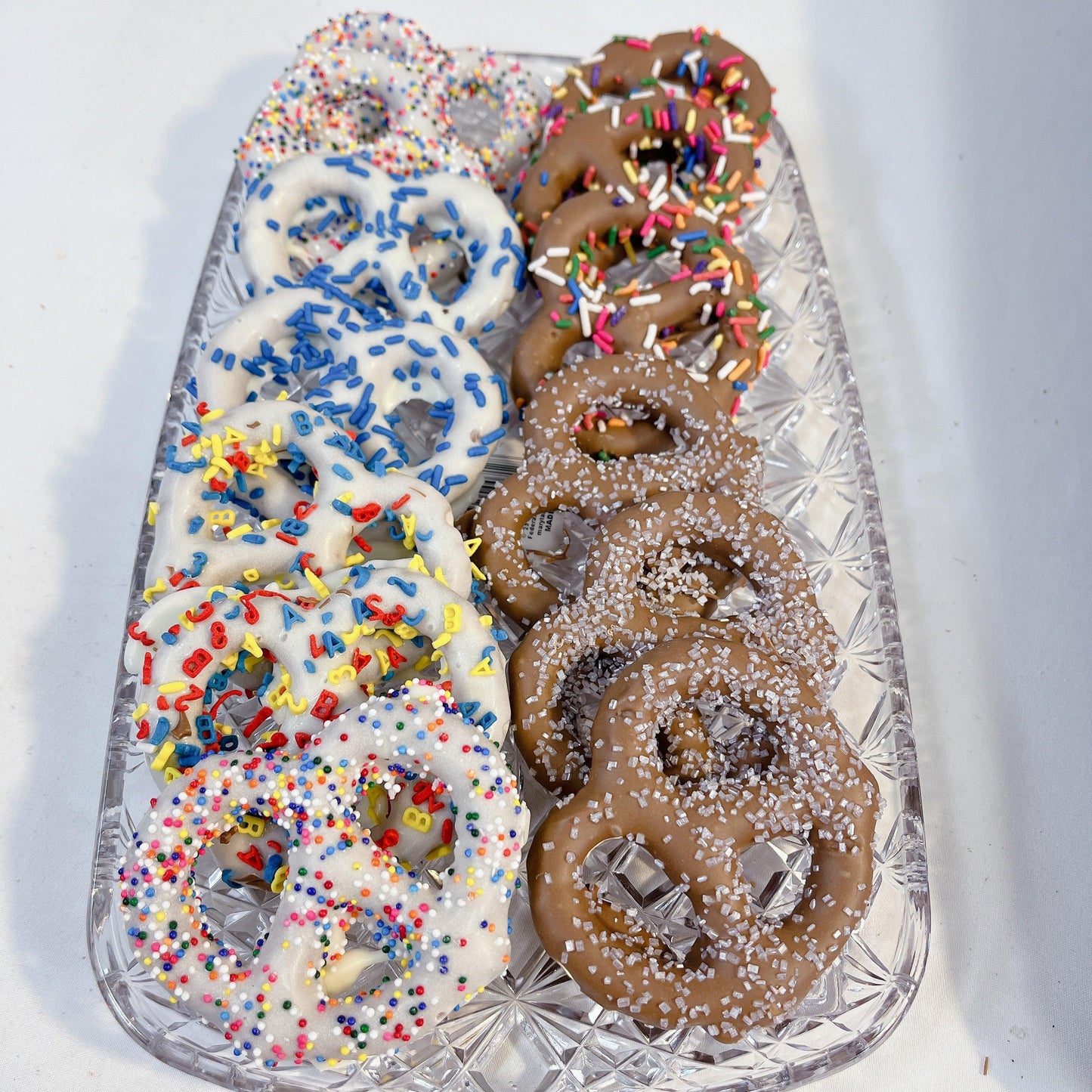 Gourmet Chocolate Dipped Pretzels with rainbow and letter sprinkles on a crystal tray. A crunchy sweet and salty treat for chocolate lovers.