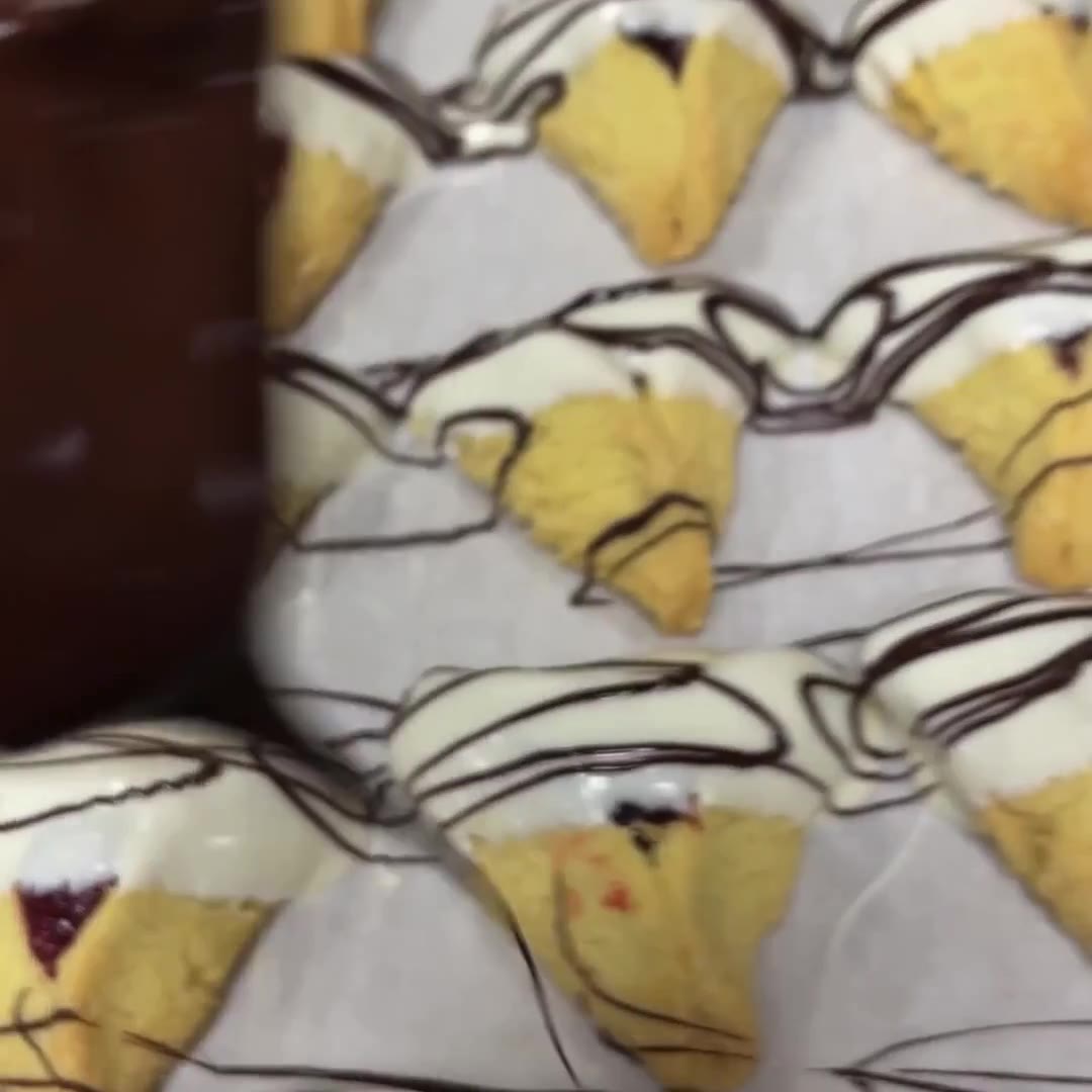 Freshly baked hamantaschen cookies dipped in white chocolate and drizzled with dark chocolate, cooling on a parchment-lined tray. Perfect for Purim celebrations.